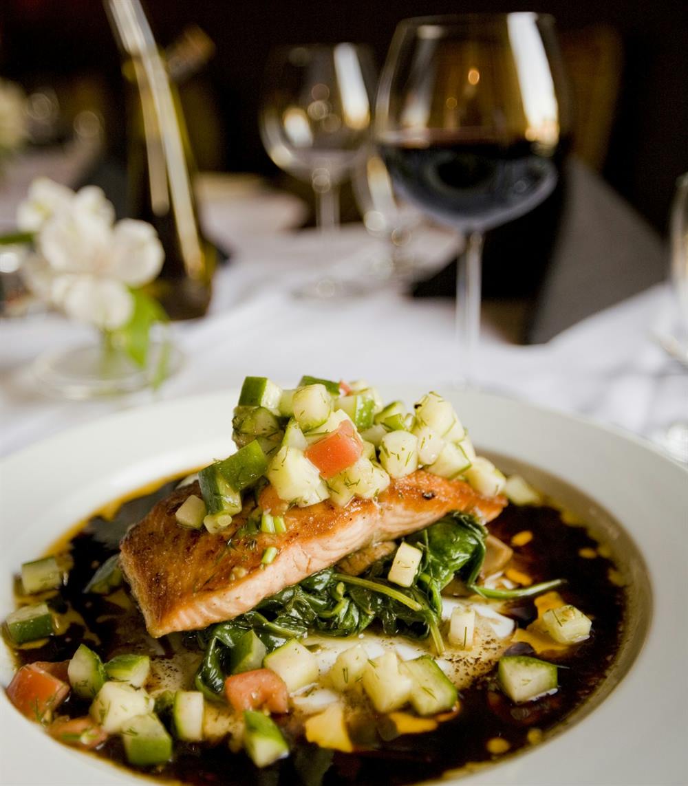 A plated salmon dish on a white table, with wine glasses beyond.